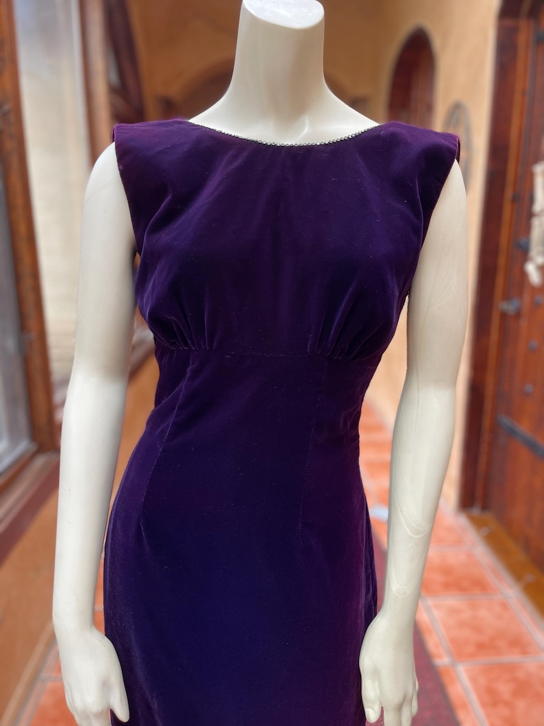 May include: A sleeveless, floor-length, deep purple velvet dress. The dress has a scoop neckline with a delicate trim and a fitted bodice that flares out at the waist. The dress is displayed on a mannequin.