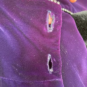 May include: Close-up of a deep purple velvet garment with two buttonholes. The edge is trimmed with a row of small, sparkling rhinestones. The fabric shows signs of wear and age.