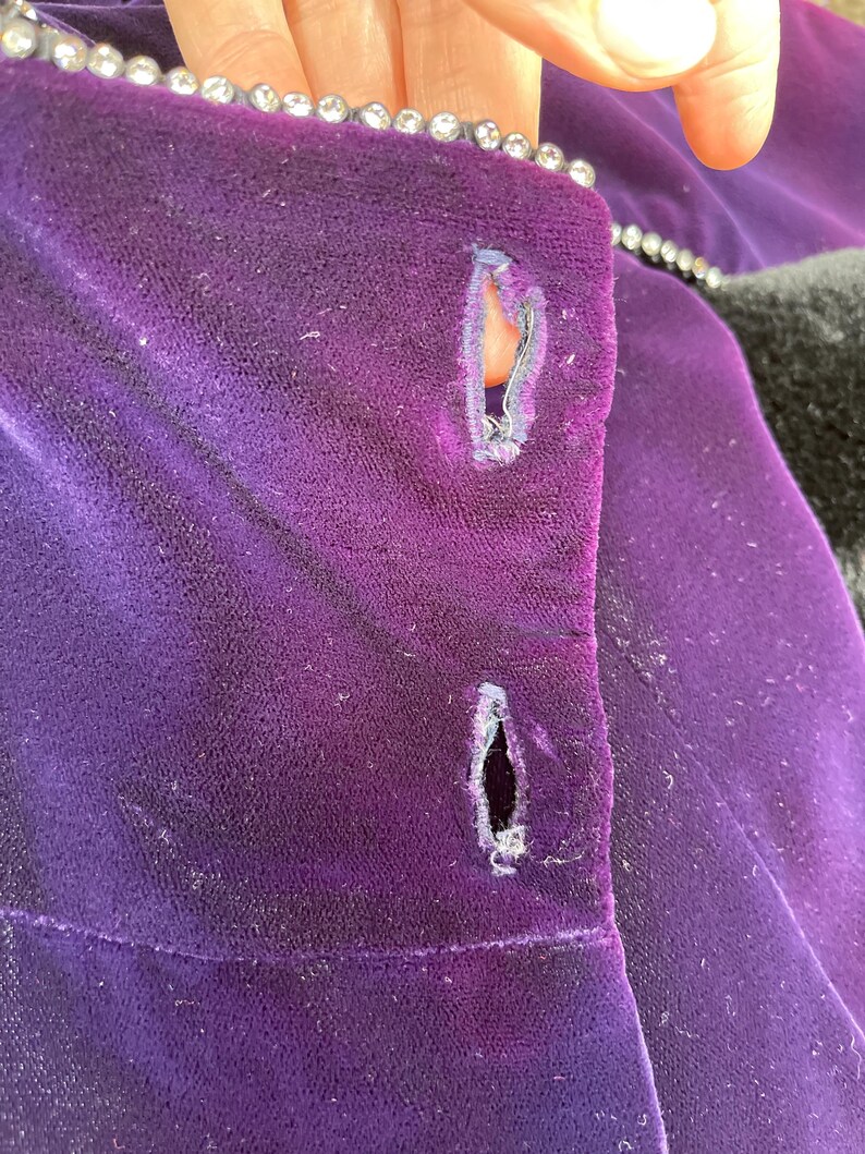 May include: Close-up of a deep purple velvet garment with two buttonholes. The edge is trimmed with a row of small, sparkling rhinestones. The fabric shows signs of wear and age.