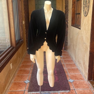 Puede incluir: Una chaqueta de terciopelo negro con escote en V profundo, mangas largas con botones dorados en los puños y un dobladillo festoneado. La prenda se cierra en la parte delantera con dos botones dorados, ofreciendo un aspecto estructurado.