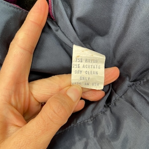 May include: A close-up of a clothing label with the text "75% RAYON 25% ACETATE DRY CLEAN ONLY MADE IN USA". The label is white with black text, set against a dark blue fabric with a magenta trim.