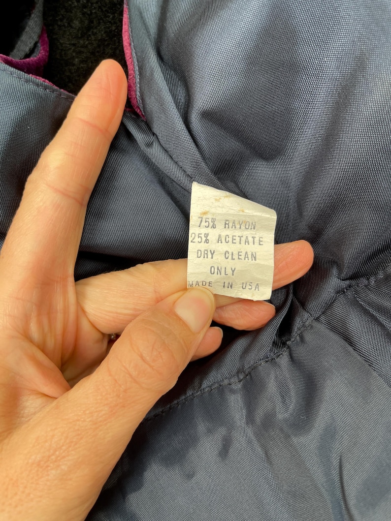 May include: A close-up of a clothing label with the text "75% RAYON 25% ACETATE DRY CLEAN ONLY MADE IN USA". The label is white with black text, set against a dark blue fabric with a magenta trim.