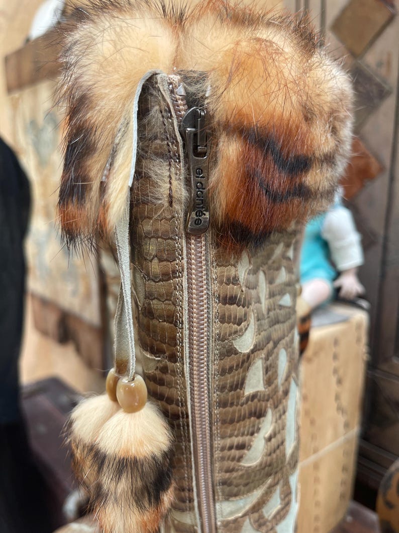 May include: Close-up of a brown and tan leather boot with a fur trim. The boot features a zipper with the text "el dante" and a decorative tassel. The boot has a snakeskin pattern with cut-out designs.