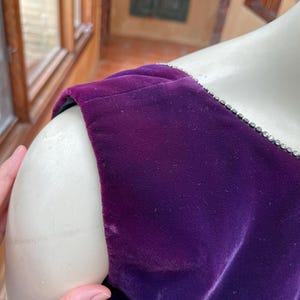 May include: Close-up of a deep purple velvet dress shoulder. The dress has a jewel-encrusted trim along the neckline. The fabric appears soft and luxurious. The dress is displayed on a mannequin.