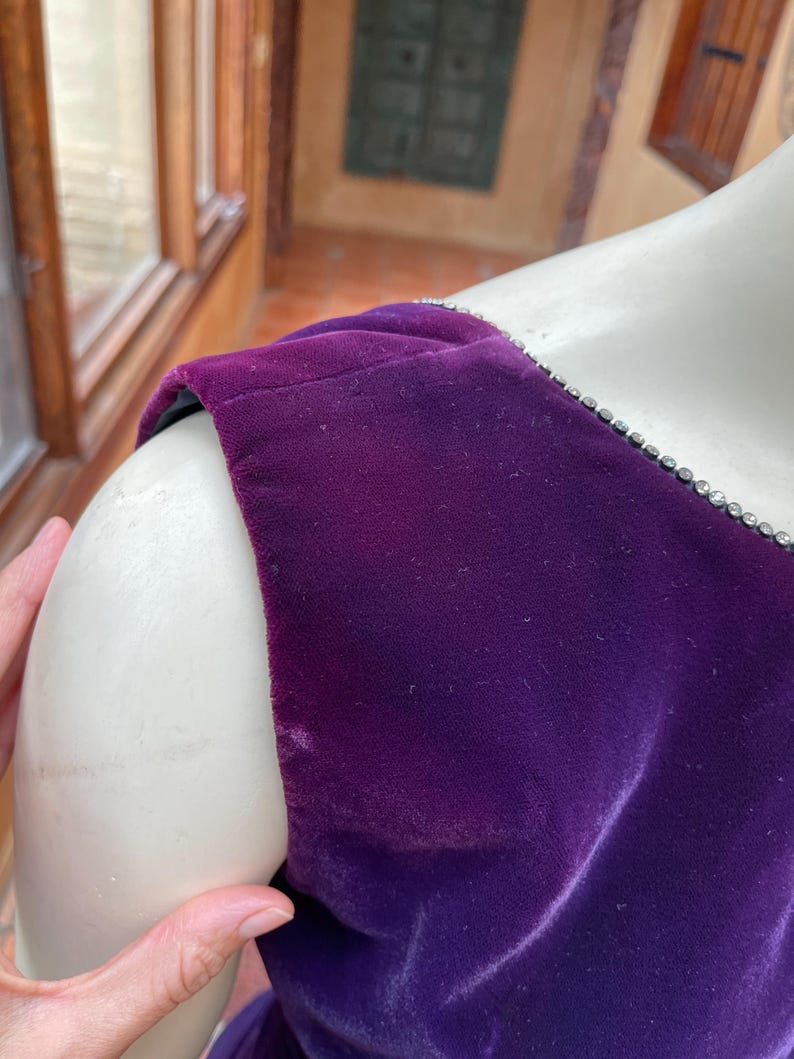 May include: Close-up of a deep purple velvet dress shoulder. The dress has a jewel-encrusted trim along the neckline. The fabric appears soft and luxurious. The dress is displayed on a mannequin.