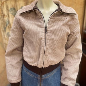 May include: A beige corduroy jacket featuring a full-length front zipper, a pointed collar, and dark brown ribbed knit cuffs and hem. The jacket has long sleeves and a textured vertical striped pattern across the fabric, worn over blue jeans.