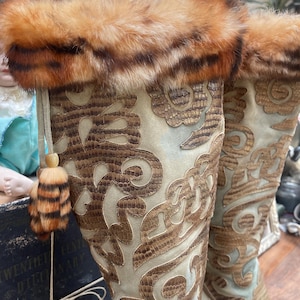 May include: A pair of tall, ornate boots with a light blue suede-like base and intricate brown leather-like detailing. The boots feature a fur trim at the top and a decorative tassel. The design is reminiscent of vintage or historical fashion.