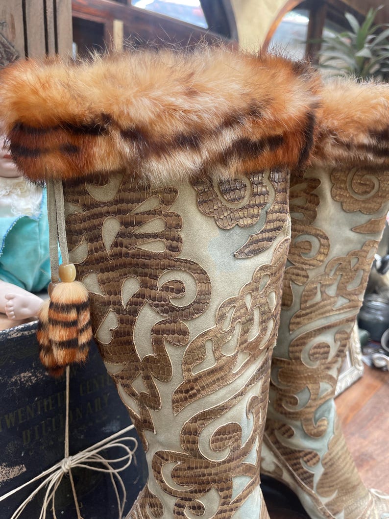 May include: A pair of tall, ornate boots with a light blue suede-like base and intricate brown leather-like detailing. The boots feature a fur trim at the top and a decorative tassel. The design is reminiscent of vintage or historical fashion.