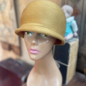 May include: A mustard yellow felt cloche hat with a rounded crown and a narrow, slightly upturned brim. The hat features a matching thin grosgrain ribbon band encircling the base of the crown, with a vibrant red lining visible underneath the brim.