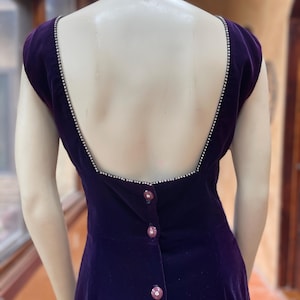 May include: A dark purple velvet dress with a low back and cap sleeves. The neckline and back are trimmed with small, sparkling embellishments. The dress has decorative buttons down the back.