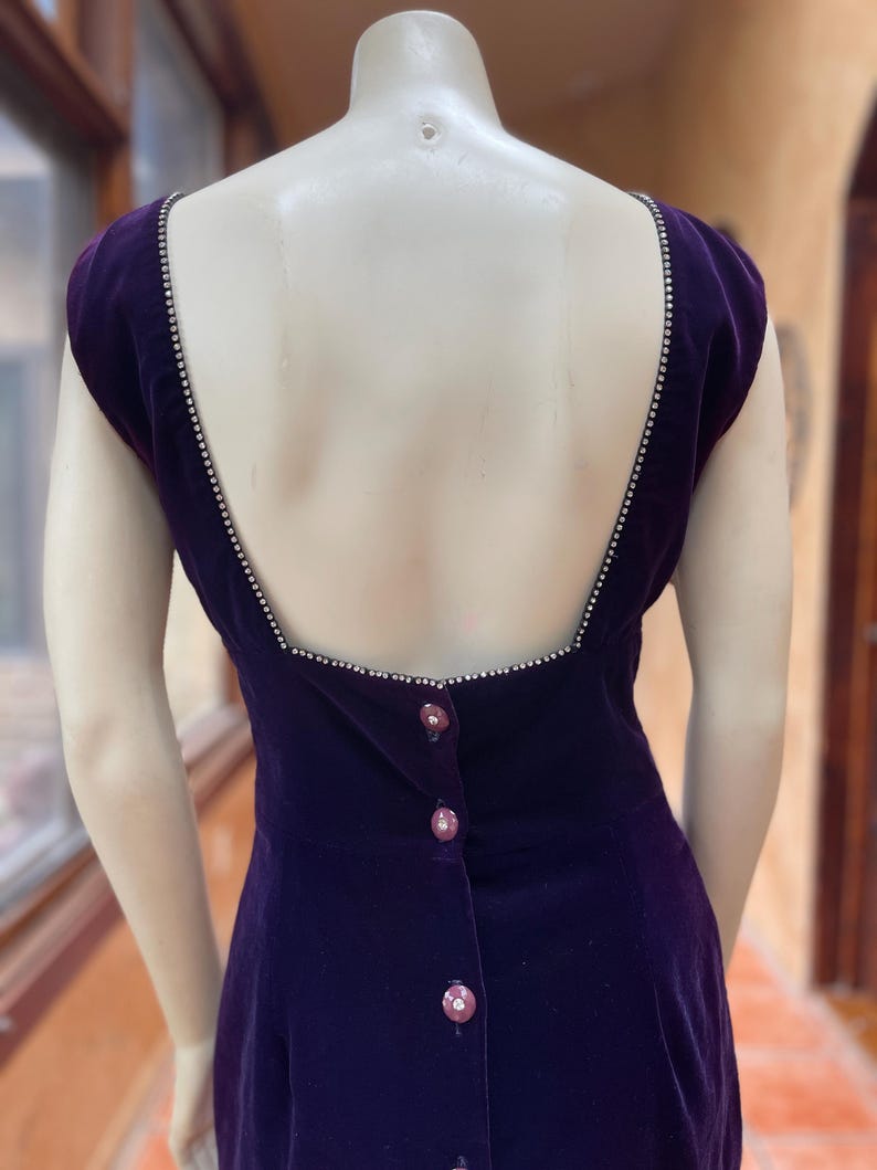 May include: A dark purple velvet dress with a low back and cap sleeves. The neckline and back are trimmed with small, sparkling embellishments. The dress has decorative buttons down the back.