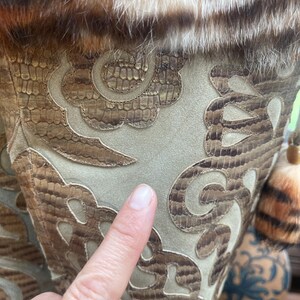 May include: Close-up of a decorative vase with a faux fur trim and intricate patterned design. The vase features a snakeskin-like pattern in shades of brown and tan, with a light green suede-like background. A finger points to the vase.