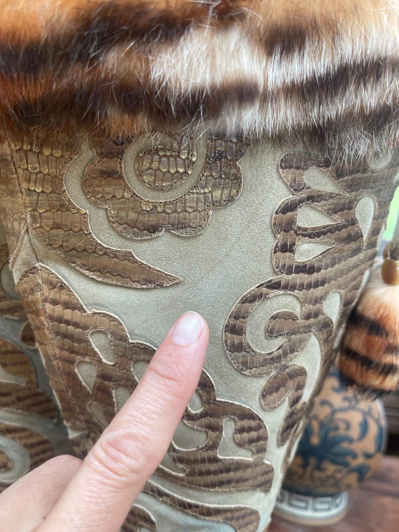 May include: Close-up of a decorative vase with a faux fur trim and intricate patterned design. The vase features a snakeskin-like pattern in shades of brown and tan, with a light green suede-like background. A finger points to the vase.