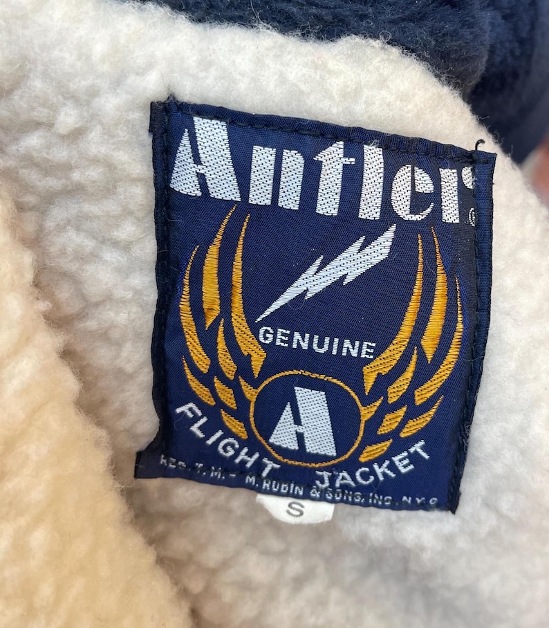 May include: A blue and yellow patch with the text "Antler Genuine Flight Jacket" and a lightning bolt graphic. The patch has a white border and the text "Reg. T.M. - M. Rubin & Sons, Inc. N.Y.C." is printed below the main text.