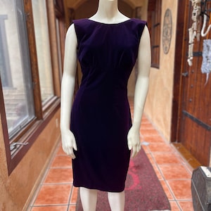 May include: A dark purple velvet dress, sleeveless and knee-length, displayed on a mannequin. The dress features a fitted bodice and a straight skirt. The mannequin stands on a metal stand in a room with windows and a patterned rug.