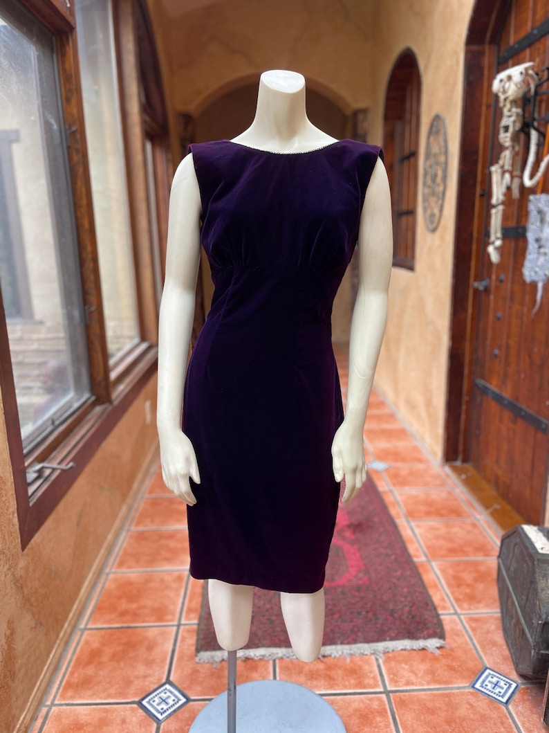 May include: A dark purple velvet dress, sleeveless and knee-length, displayed on a mannequin. The dress features a fitted bodice and a straight skirt. The mannequin stands on a metal stand in a room with windows and a patterned rug.