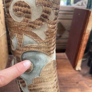 May include: Close-up of a tall boot with a snakeskin pattern and decorative cutouts. The boot features a mix of tan, brown, and light green suede-like materials. The design includes intricate, swirling patterns.