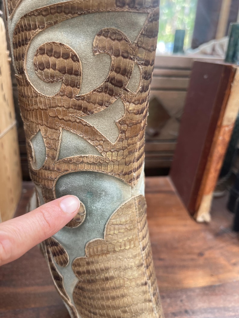 May include: Close-up of a tall boot with a snakeskin pattern and decorative cutouts. The boot features a mix of tan, brown, and light green suede-like materials. The design includes intricate, swirling patterns.
