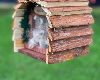 May include: A small wooden birdhouse with a brown and tan finish. The birdhouse is shaped like a cabin and has a small opening for birds to enter. It is hanging from a wire.