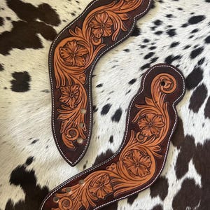May include: A pair of curved leather spur straps featuring intricate floral tooling and scrollwork. The leather is dyed in a deep brown tone with natural tan highlights in the embossed flower patterns, finished with white stitching along the edges.
