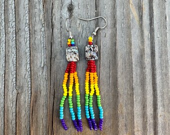 May include: Handmade beaded earrings with a rainbow color scheme. The earrings feature square, patterned beads at the top, with strands of red, orange, yellow, green, blue, and purple beads hanging below. The earrings are displayed on a weathered wooden surface.