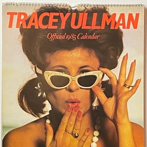 May include: A vintage calendar featuring a woman with dark hair wearing white cat-eye sunglasses and a white pearl necklace. The calendar is titled "Tracey Ullman Official 1985 Calendar."
