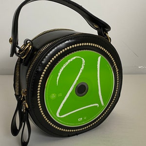 May include: A black round purse with a gold zipper and a gold trim. The front of the purse has a green CD with the number 21 written on it.
