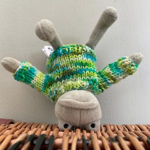 Op de afbeelding: Een pluche kikker met grijze ledematen, gekleed in een groen, blauw en geel gebreide trui. De kikker ligt ondersteboven op een geweven oppervlak. Het speelgoed heeft zwarte ogen en een lichtbruine kop.