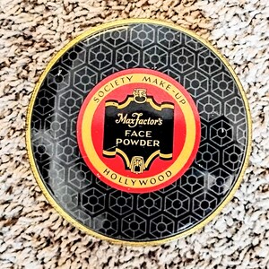 Könnte beinhalten: Vintage Max Factor's Face Powder Kompaktpuder. Der runde Kompaktpuder hat ein schwarzes geometrisches Muster mit einem goldenen Rand. Das zentrale Etikett ist rot mit goldenem und schwarzem Text: "SOCIETY MAKE-UP Max Factor's FACE POWDER HOLLYWOOD."