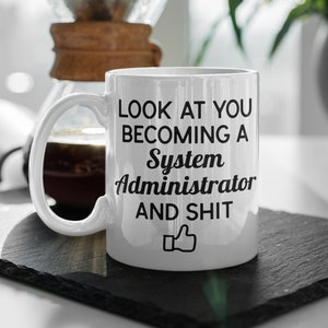 May include: White ceramic coffee mug with black text that reads "Look at you becoming a System Administrator and shit" with a thumbs up icon.