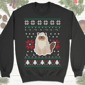 May include: A black Christmas sweater with a white, green, and red snowflake pattern. A white cat with brown eyes and a brown tail is in the center of the sweater.
