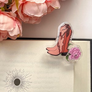 May include: A clear acrylic bookmark featuring a pink and coral cowboy boot design with brown accents and a pink flower. The bookmark is placed on the pages of a book, with a sunburst design visible on the page.