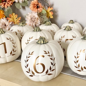 May include: Nine white pumpkins with gold leaf numbers 1 through 7, and 2, 5, and 6. Each pumpkin has a gold leaf wreath around the number.