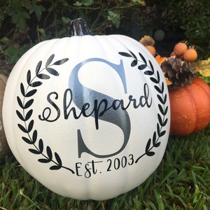 May include: White pumpkin with black lettering that reads "Shepard" with a large "S" in the center, surrounded by a wreath. The text "Est. 2003" is below. A small orange pumpkin and other fall decor are in the background.