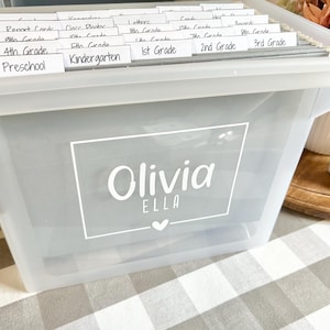 May include: A clear plastic storage bin with white labels for different grade levels, from preschool to 3rd grade. The bin is labeled "Olivia Ella" with a heart.