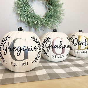 May include: Three white pumpkins with black stems, personalized with names and dates. Each pumpkin features a different name in a decorative font, surrounded by a black leaf wreath. The pumpkins are on a gray and white checkered table runner.