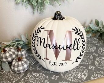 May include: White pumpkin with a black and rose gold monogram "M" with the text "Maxwell Est. 2019" on the front. The pumpkin is sitting on a gray and white patterned surface.