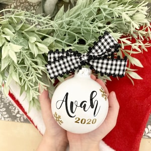 May include: White Christmas ornament with black and white checkered ribbon and gold snowflakes. The ornament is personalized with the name "Ava" and the year "2020".