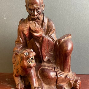 Peut inclure: Une sculpture en bois brun foncé d'un homme assis avec une longue barbe tenant un livre. Un animal en bois sculpté plus petit avec une expression de grognement est assis aux pieds de l'homme.