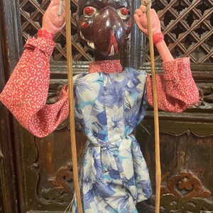 May include: A wooden puppet with a carved, painted face and a floral patterned dress. The puppet is holding two wooden sticks and is standing on a blue platform.