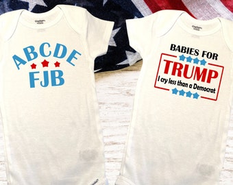 May include: Two white baby onesies with political slogans printed on them. The onesie on the left says "ABCDE FJB" in blue letters with red stars. The onesie on the right says "Babies for Trump I cry less than a Democrat" in red, blue, and white letters.