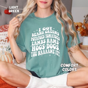 May include: A light green t-shirt with a white graphic that says "I got beans, greens, potatoes, tomatoes, lambs, rams, hogs, dogs, you name it!" The shirt is worn by a person sitting on a couch. The text "Comfort Colors" is visible in the bottom right corner of the image.