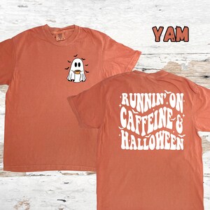 May include: A burnt orange t-shirt with a white ghost holding a cup of coffee on the front and the text "Runnin' on Caffeine & Halloween" on the back.