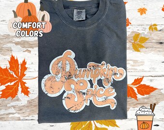 May include: A dark grey t-shirt with a distressed white and orange graphic that says "Pumpkin Spice". The shirt is folded and laid on a wooden surface with autumn leaves and pumpkins.