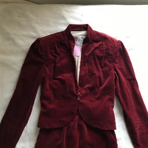 May include: A burgundy velvet blazer with a mandarin collar and a single button closure. The blazer has a fitted silhouette and long sleeves.