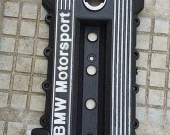 May include: Black engine valve cover with silver "BMW Motorsport" lettering and multiple circular openings.