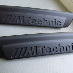 以下が含まれることがあります： 「M Technic」という文字がエンボス加工された黒いプラスチック製のトリムが2つあります。