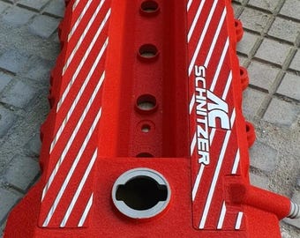 Può includere: Un coperchio valvole motore rosso con strisce diagonali bianche e la scritta "AC Schnitzer". Il coperchio ha diversi fori e una superficie strutturata. Progettato per uso automobilistico.
