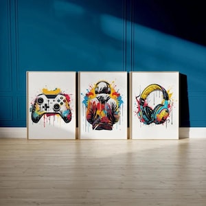 Pode incluir: Um conjunto de três quadros de parede emoldurados com designs vibrantes de salpicos de tinta multicoloridos. O quadro da esquerda mostra um comando de videojogos, o central uma pessoa com capuz e auscultadores, e o da direita auscultadores.
