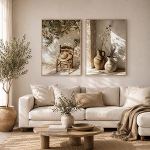 May include: Two framed wall art prints displayed above a beige sofa. The left print shows a wooden chair with a straw hat under olive branches. The right print features two ceramic vases with dried flowers, both bathed in warm, natural sunlight.